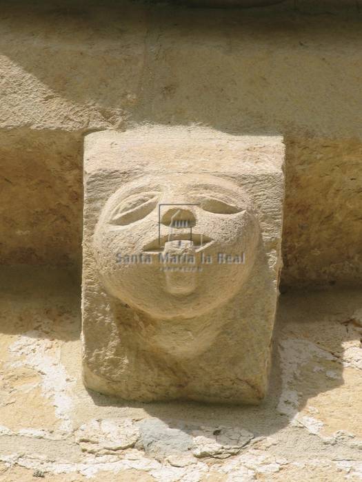 Canecillo esculpido de cabeza humana redondeada que saca la lengua,encima de la portada en el muro de la epistola