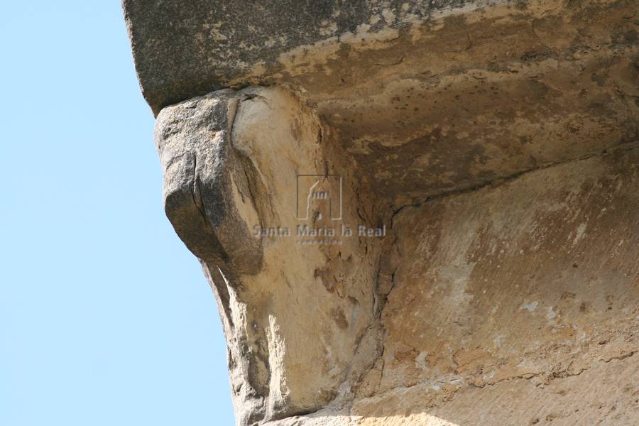 Canecillo en el muro norte de personaje en cuclillas que se levanta la falda sobre las rodillas ,muy deteriorado