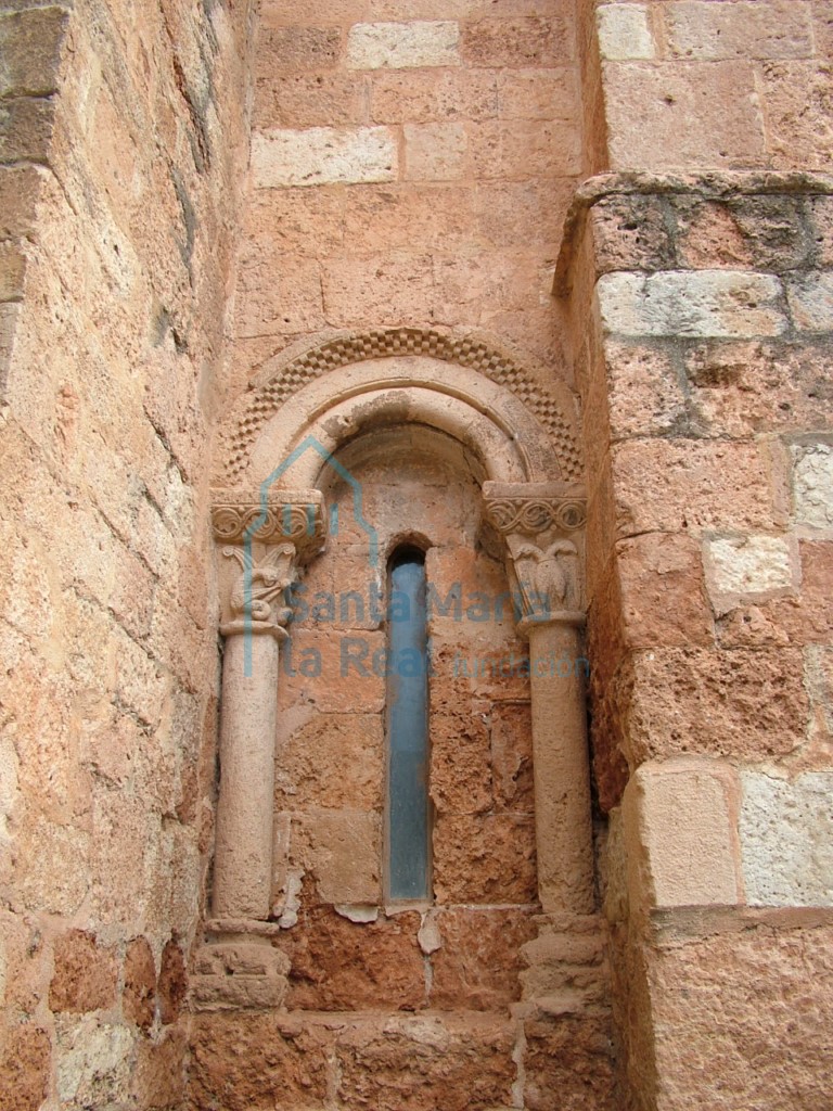 Aspillera del frente sur del tramo recto de la cabecera, cuenta con un arco de doble medio punto de ancho bocel y ajedrezada chambrana apoyado en dos columnillas de capiteles decorados con iconografía animal