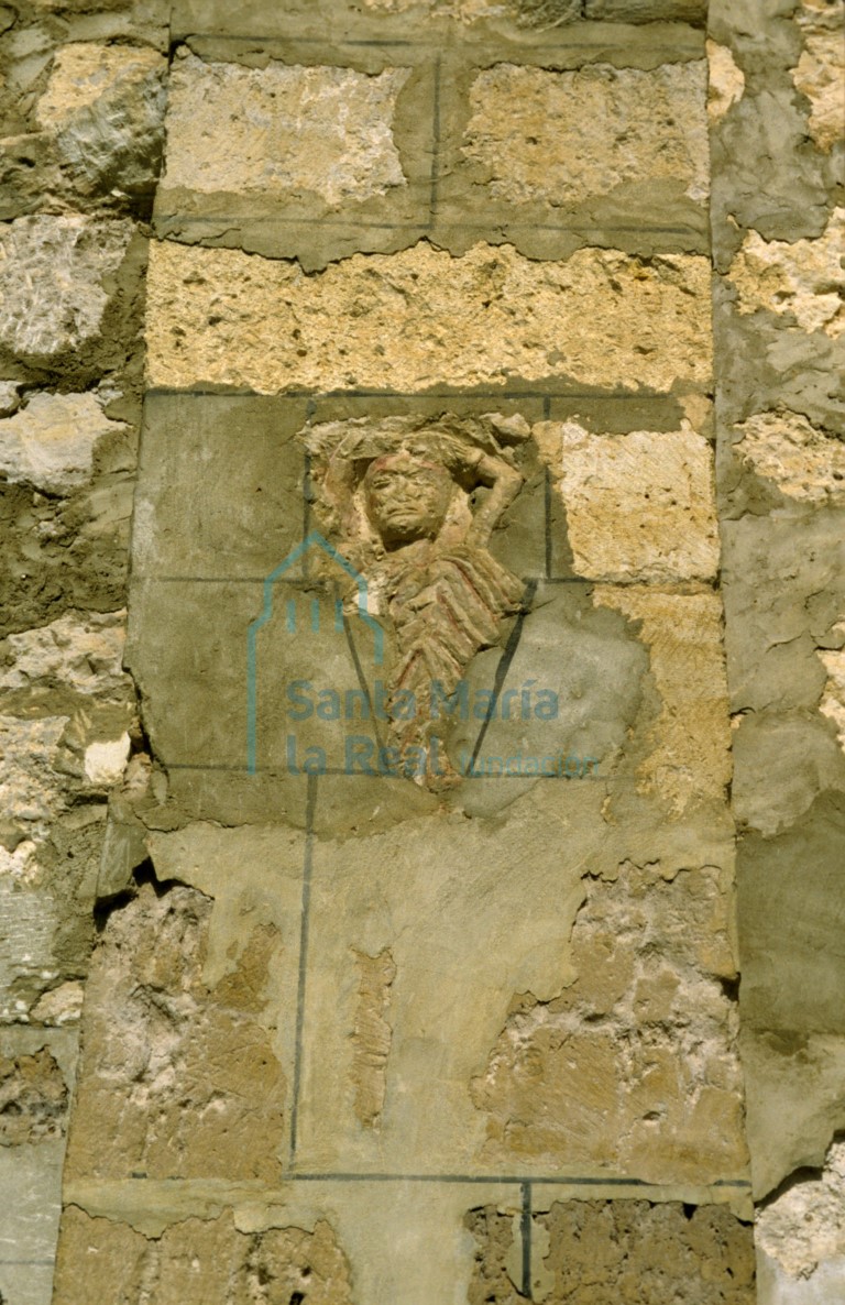 Relieve engastado en un contrafuerte del muro sur. Personaje masculino que alza sus brazos hacia su testa, viste manto de acusados pliegues triangulares